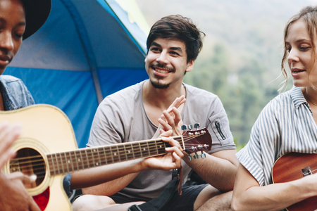 Group of young adult friends in camp site playing guitar and ukelele and singing together outdoors...
