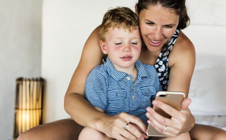 Mother sitting with son on bed using mobile