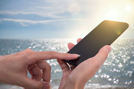 Close up of a woman using mobile smartphone outdoor at sand beach with sparkling sea waterの写真素材