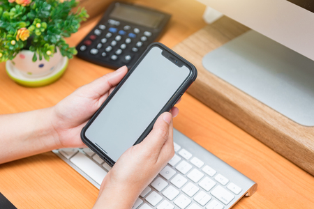 Desk in the office Hand is holding showing smartphone with blank mock up screen over office desk table. This can be used to montage the mobile application.の写真素材