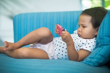 Chubby baby using a pink cell phone by herself on the sofaの写真素材