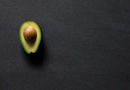 half of a avocado still with the stone on a dark black textured background from above side lighting