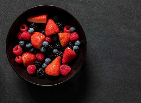 strawberries raspberries blackberries and blue berries in a black bowl with a textured background...