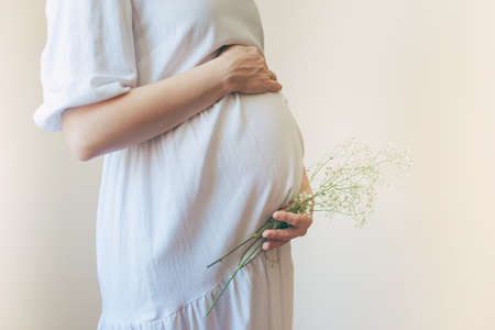 Pregnant woman in white dress holding belly and flowers with her hands.