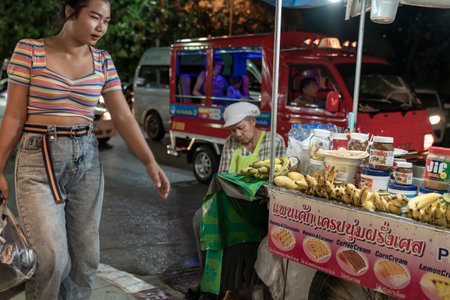 Thailand, Phuket, Patong, February 1, 2020: Asian Thai woman walks past street food banana pancakes point, night street photo, blurr, soft focus,のeditorial素材