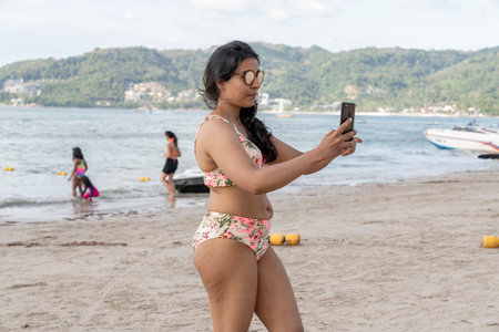 Thailand, Phuket, March 30, 2020: an Arab or Eastern Indian woman with sunglasses takes photos of herself . Selfie on the beach in a swimsuit. cellulite on the thighs and stomachのeditorial素材