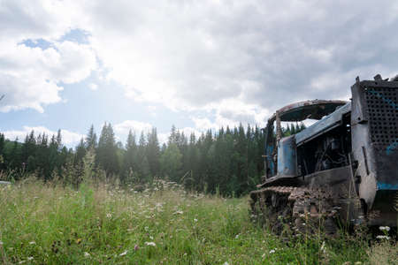 an old rusty crawler tractor, abandoned in a field, against the background of a pine forest in Siberia. decline and collapse of agriculture in Russiaの写真素材