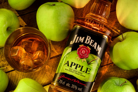 Still life with a bottle of Jim Beam Apple Bourbon (Apple liqueur from Kentucky) with a glass of cold whiskey on ice on a wooden brown background of boards, surrounded by juicy green apples.のeditorial素材