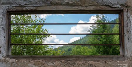 an old window with rusty bars and a beautiful view of the green forest and cloudy sky. Hope for the...