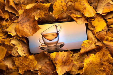 a mug of tea on the mobile phone screen against the background of autumn leaves. orange cocept...