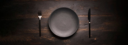 A black empty plate and a fork with a knife on a dark background. kitchen restaurant invitation event, panoramic photoの写真素材