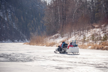 Winter walk with family on a snowmobile on a frozen river. A snowmobile with a trailer rolls children on the ice of the frozen Siberian Mana River in early spring.の写真素材