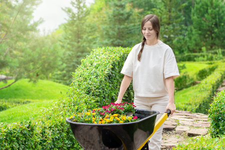 A woman tends to her garden, moving a wheelbarrow filled with colorful flowers through lush greenery.の写真素材