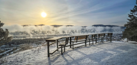 Tranquil winter scene with sun shining over misty valley, snowy ridge, bench, breathtaking viewの写真素材