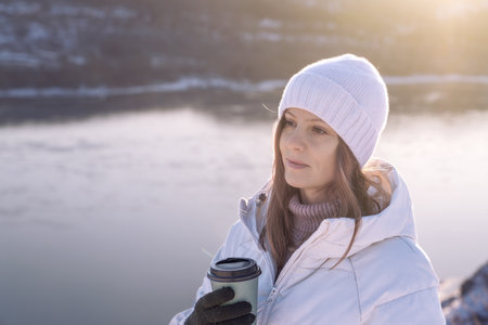 A woman in a white jacket and hat drinks hot coffee in winter by the riverbank and enjoys the moment and silence. A Winter Woman Happily Enjoying a Warm Beverage While Relaxing Near a Beautiful Frozen Lakeの写真素材