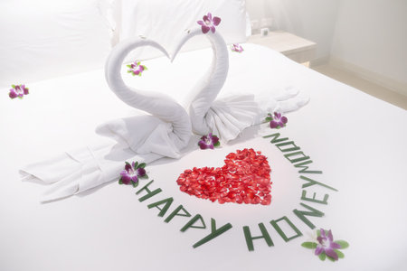The honeymoon. Two beautiful snow-white swans, as a symbol of love and relationships, decorated with orchid flowers on a white bed for lovers close-up. The Happy Honeymoon inscription is lined with green plant stems.の写真素材