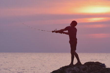 Sea Fishing. A silhouette of a fisherman casts a spinning rod from a rocky cliff into the Andaman...