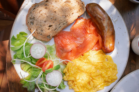 Breakfast platter featuring smoked salmon, scrambled eggs, sausage, vegetables, and whole grain bread served on a sunny morning at a local cafe close-up, top viewの写真素材