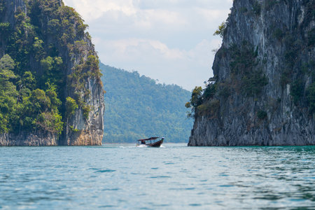 Small boat glides across the turquoise waters of Cheow Lan Lake, by towering limestone cliffs...