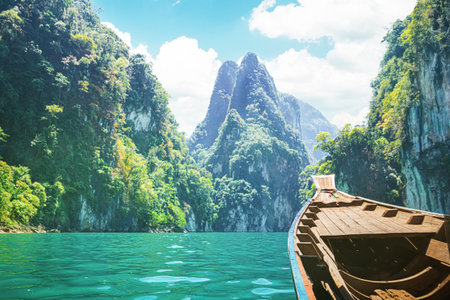 Travelers enjoy a cruise on a traditional longtail boat in Cheow Lan Lake, surrounded by towering limestone cliffs and lush greenery. The vibrant blue waters and scenic mountainsの写真素材
