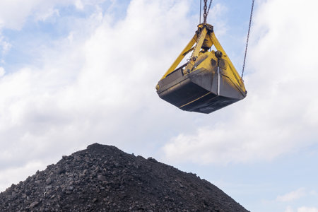 Heavy-duty coal bucket being unloaded by an excavator crane above a large pile of coal at a mining site, Coal delivery and shipment to consumers, coal pricesの写真素材