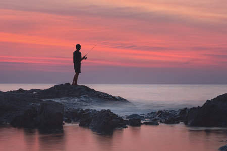 A striking silhouette fisherman, is seen at sunset over a rocky shoreline with gentle and calm ocean...