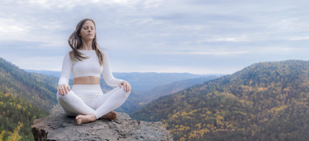 White woman meditates in lotus position on a stone cliff surrounded by autumn mountains in nature. A young woman practices yoga and fitness outdoor copy spaceの写真素材