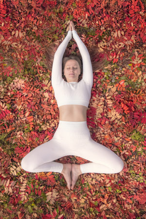 A woman is engaged in a yoga pose amidst a vibrant carpet of autumn leaves. Her white attire contrasts beautifully with the rich reds and oranges of the foliage.の写真素材