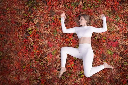 In a tranquil outdoor space adorned with colorful autumn leaves, a woman in white yoga attire performs a pose on the ground. red background copyspaceの写真素材