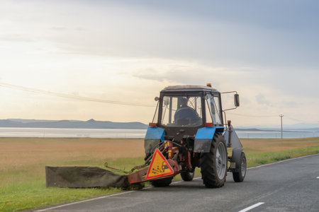 Tractor cutting grass along an empty rural road near a serene lake during a cloudy afternoon. Roadside work, mowing grass along roads and highwaysの写真素材