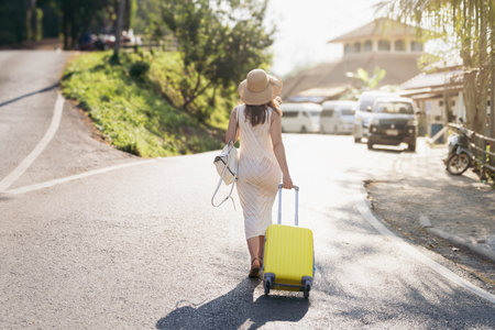 A Young Traveler Carrying a Bright Yellow Suitcase on a Beautiful Scenic Road in a Wonderful Sunny Destinationの写真素材
