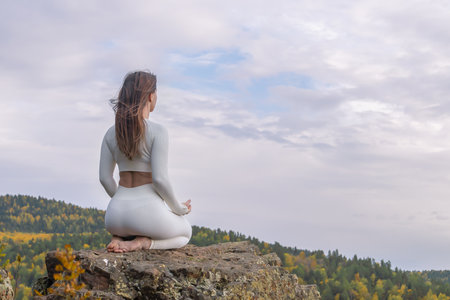 Yoga and zen. A woman sits gracefully on a rocky surface atop a mountain, dressed in white clothing. She is in a meditation pose, gazing into the distance amidst the colorful autumn foliage.の写真素材