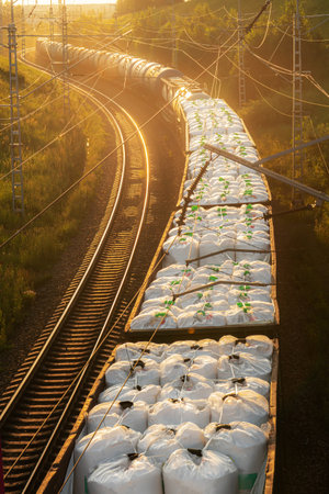 A train with wagons loaded with chemicals or mineral fertilizers in bales and bags travels at sunset along the railway tracks. Fertilizer delivery and food security.の写真素材