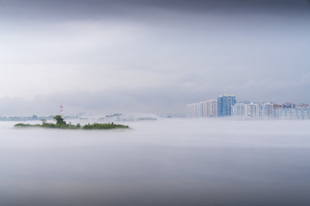 Fog blankets the Yenisei River, creating a mystical atmosphere in the residential area of Krasnoyarsk. New buildings rise above the mist, contrasting with the serene water and lush green islands.の写真素材
