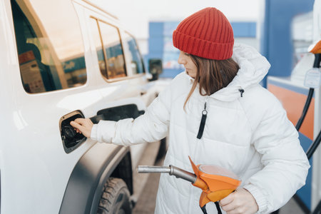 A woman in a white down jacket refueling her car at a gas station. Winter car trips and fuel prices. her vehicle at a gas station during winter car trip while managing fuel costsの写真素材