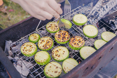 Fresh zucchini and eggplant slices are being grilled on a metal rack placed over glowing coals. A hand carefully flips the vegetable slices to achieve an even roast.の写真素材
