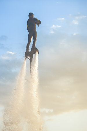 Person practicing flyboarding at sunset in an open sky above water with beautiful clouds. Mans...