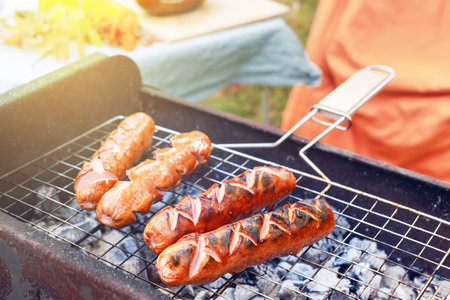 Grilled sausages are sizzling on a barbecue grill filled with glowing coals. The scene captures the essence of an autumn picnic, with warm sunlight filtering through.の写真素材