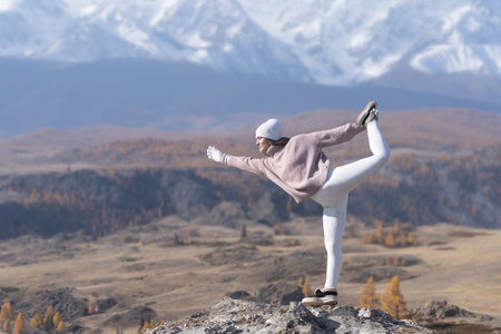 A woman balances on one leg on a rocky ledge high in the mountains. Dressed in warm, cozy activewear, she enjoys the fresh air and vibrant autumn colors of nature while radiating wellness.の写真素材