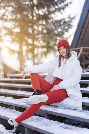 Young woman in winter fashion lounging on snowy steps in a sunny outdoor setting during daylightの写真素材