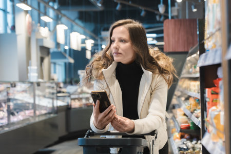 In a modern supermarket, a woman pushes a grocery cart while looking at her smartphone, recalling...