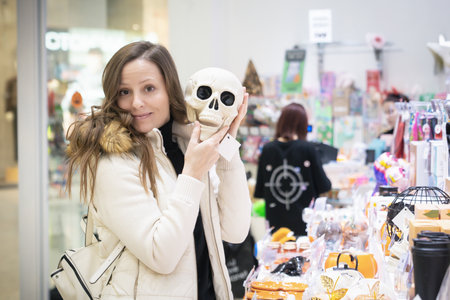 A white woman is smiling while holding a plastic toy skull in a shopping center filled with Halloween decorations and gifts. She is enjoying the fun atmosphere of holiday shopping.の写真素材