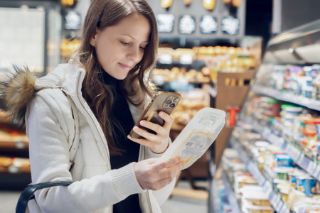 A woman in a coat uses her phone to scan a cheese package label while shopping in the grocery store. She checks the ingredients and expiration information before making a purchaseの写真素材