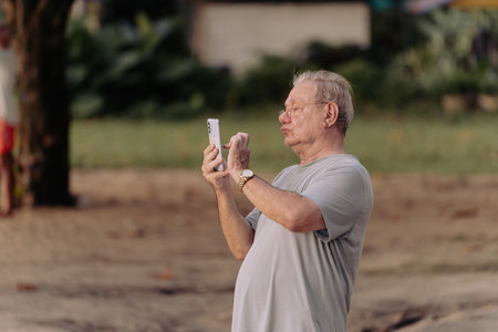 Khao Lak, Thailand, 24 11 2025: an elderly man with glasses on vacation by the sea takes pictures of the sunset on his smartphone, side view close-up portrait lifestyle. High quality photoのeditorial素材