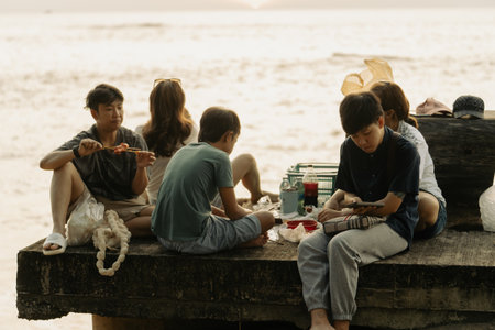 Khao Lak, Thailand, 24 11 2025: real authentic moments of life- A group of young Thai teenagers on a picnic near the sea in summer on the Khao Lak embankment eating kebabs and enjoying the sunsetのeditorial素材