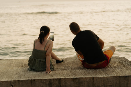Khao Lak, Thailand, 24 11 2025: real authentic moments of life- A couple in love, a young family photographing the sunset by sea, sitting on the embankment back view, real moments vacation lifestyleのeditorial素材