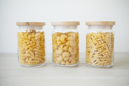 three transparent pots with wooden tops full of different kinds of raw pasta on light grey background. selective focus. shallow depth of field.の写真素材