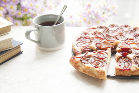 Whole plum cake with tea mug, books and flowers on a wooden background. Soft warm daylight. Selective focus.の写真素材