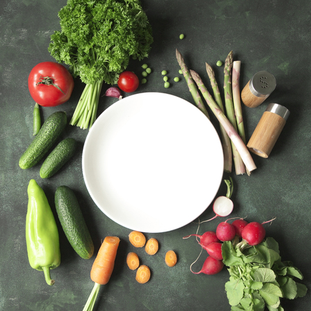 A white plate surrounded by a variety of fresh raw vegetables on a rustic dark green background. Food frame.の写真素材