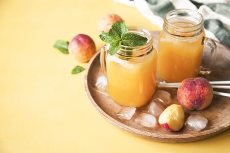 Cold summer peach drink with ice and mint in mason jars on a tray with fresh peaches on yellow background. Copy space.の写真素材